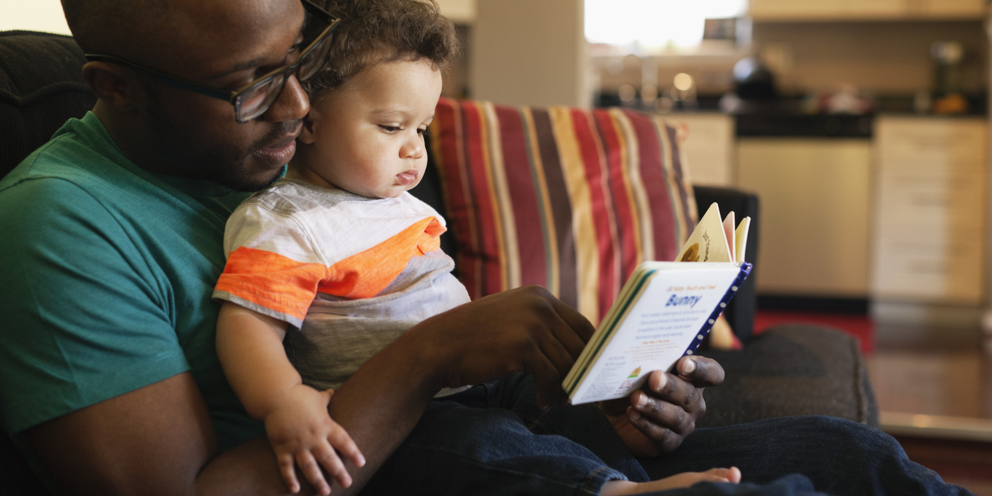  A father sits on a couch and reads a book to his baby.