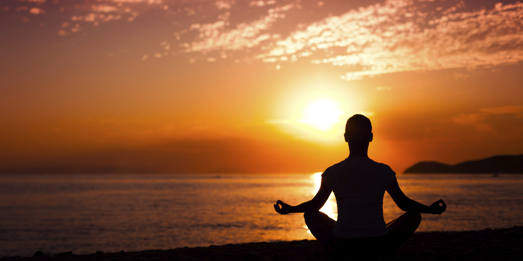  A person sits on a beach in a meditative pose as the sun sets over the ocean.