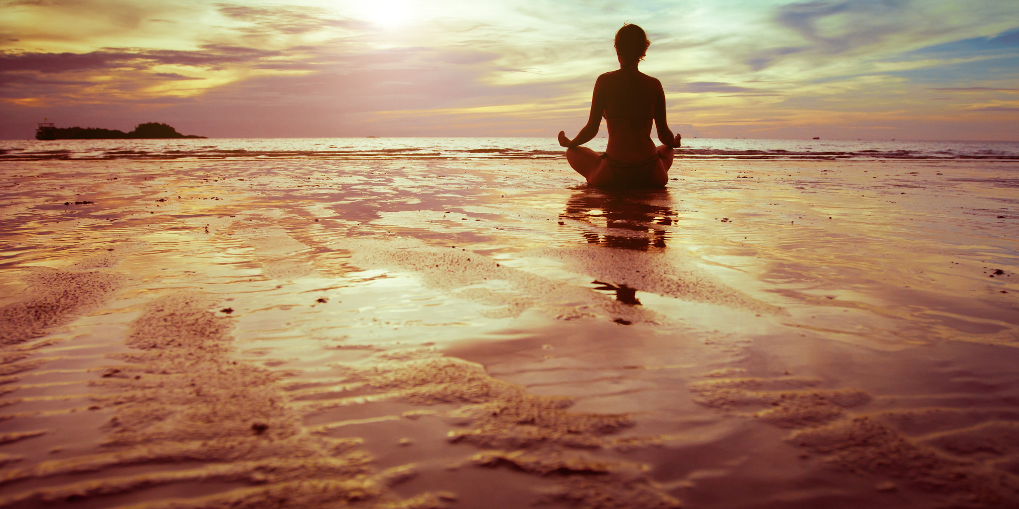  A person sits in meditation pose on a beach with the ocean and sky in the background.